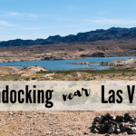 A photo of gravelly ground dotted with shrubs and a rock fire pit in front. The background shows a bright blue lake surrounded by layered (sedimentary) hills with mountains behind them. Text reads "Boondocking near Las Vegas."