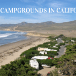 Photo of several RVs nestled between lush green landscaping the offers excellent privacy. They are just off an ocean beach with light brown hills in the background and blue sky above. Text says "Best campgrounds in California."