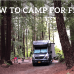 A photo of a Class B (van-style) RV in a campsite with a picnic table and camp chairs. It is surrounded by very tall cedar trees. Text says "How to camp for free."