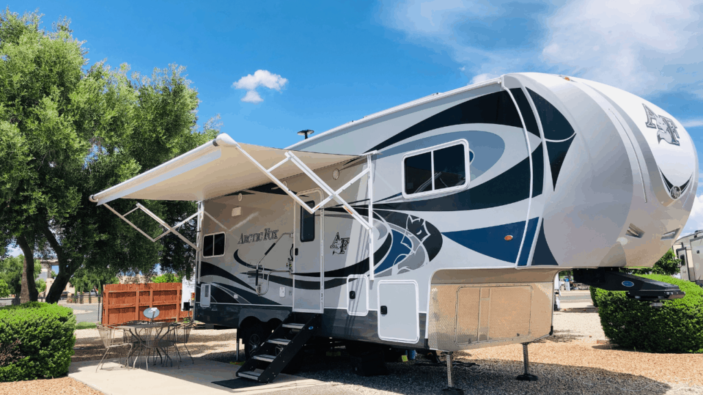 A photo of the Chickery's Arctic Fox 5th wheel with the awning out. It sits in a campsite with a paved patio and bistro-style table and chairs. The bright blue sky above has just a few wispy clouds.